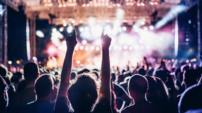 A view from behind an audience rocking out at a concert, backlit by colorful stage lighting.